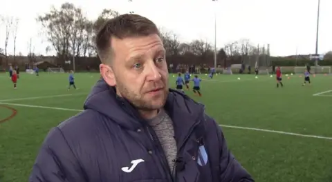 Steve Parker, chair of Crewe FC, standing in front of a pitch. He is wearing a navy blue coat.