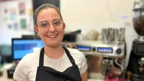 Mia Wallace is behind the counter of a coffee shop. You can see the coffee machine in the background. She is wearing a white T-shirt and black apron. 