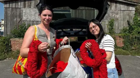 Westbury Yarn Bombers Two people stood next to each other with red poppies stood behind a car. They are smiling widely in the sunshine.