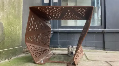 A brown bench pictured from ground level, from the side. You can see through the bench, which has solid sides with holes cut in it. It appears similar to looking through a cheese grater.