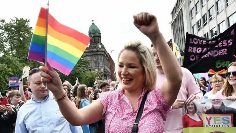 Pacemaker Michelle O'Neill has blonde hair and is wearing a pink t-shirt. She is smiling at the crowd and waving a rainbow Pride flag and pumping her fist while walking among lots of people at a Pride parade in Belfast