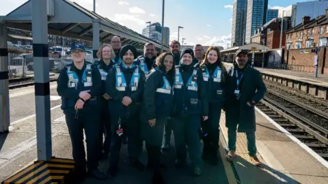 South Western Railway A group of 11 South Western Railway revenue officers, all wearing blue uniforms and blue hi vis vests, pose together on the platform of Woking station.