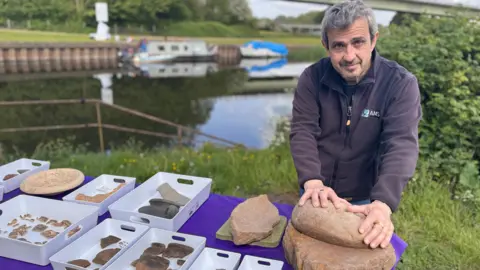 Pese Salinas from Archaeological Management Solutions, stood beside a river with a table displaying finds from the site