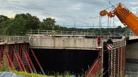 BBC A large concrete bridge structure is under construction, supported by red steel formwork and scaffolding