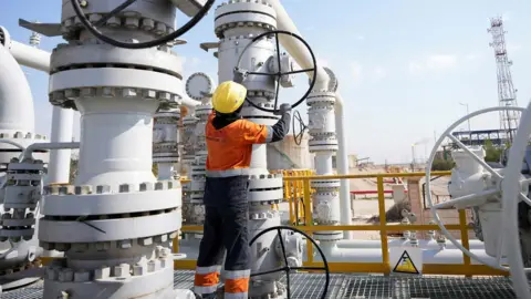 Reuters A worker operates valves at the Rumaila oil field. He is earing an organge boiler suit and yellow helmet . He is outside and there is a blue sky