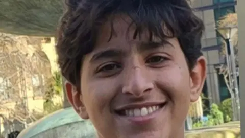 A boy smiles at the camera. He has brown hair, which falls over his forehead. The picture was taken outside, with several trees in the background.