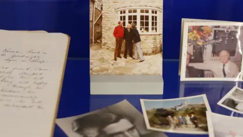 Dorset Museum A display of photographs and a notebook. The photograph in the centre shows Brynley kissing Notley on the cheek in the street while standing alongside another man.