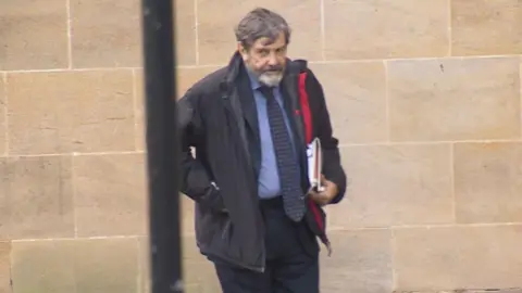BBC Alan Harris, wearing a dark coat, navy blue tie with yellow dots on it, a blue shirt and black blazer, stands next to the wall outside Winchester Crown Court where he is standing trial. A lamp-post is in the foreground of the photo.