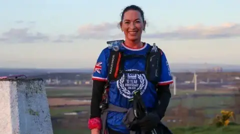 A woman wearing a blue shirt and a rucksack with attachments for water bottles is standing on top of a hill with a white-painted trig point next to her. In the distance are a series of wind turbines.