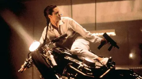 PA Media Lara Croft on a motorbike with a gun in each hand