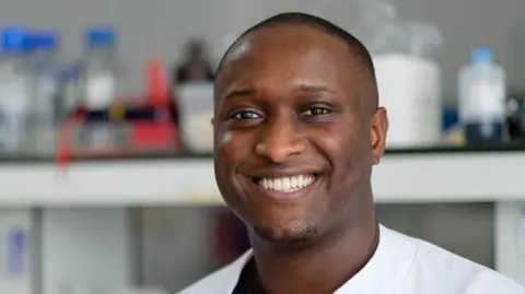 Yorkshire Cancer Research A man with a shaved head, short goatee and white shirt, in front of a blurred lab background, smiles at the camera.