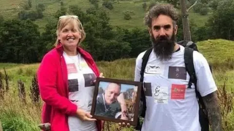 Eugene Scardifield A smiling woman in a red jacket and her adult son in a white t-shirt wearing a backpack stand in a field holding a photo of son and brother Michael who was murdered 