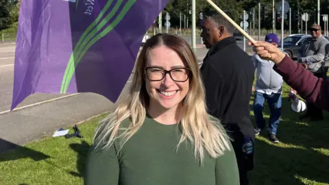 Jayne Jackson who is smiling at the camera. She has on black rimmed glasses and a green top. Behind her are people holding purple and green Unison flags. 