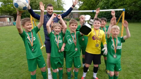 Eight boys in green football tops with medals round their necks. Two of the boys are holding up a trophy. They are in front of the goal. 