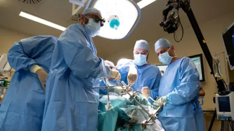 PA Media The image shows a surgical operation taking place in an operating theatre. A large circular surgical light illuminates the operating area. Several surgeons are gathered closely around a patient who is mostly covered by blue surgical drapes. 