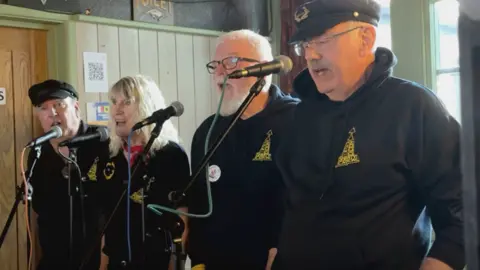 Four singers, including three men and one woman, standing on a stage and singing into microphones. They are all wearing navy jumpers with a yellow logo on the front.