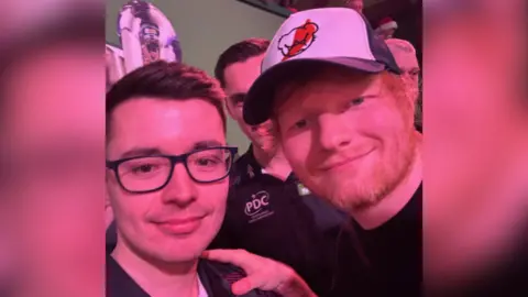 Owen Binks Ed Sheeran wearing a navy and white cap and smiling while holding Owen Binks' shoulder.