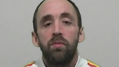 Mugshot of Mark Wallace. He has a thick dark beard and moustache and is balding with short black hair. He is wearing a white top with thick red and yellow piping on the shoulders.