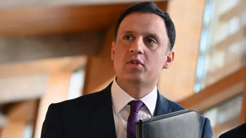 Getty Images A man with black hair walks in the Scottish Parliament. He is wearing a dark suit, shite shirt and purple tie and carrying a black folder held against his chest.