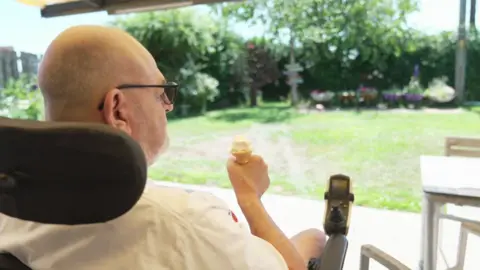 A man in a wheelchair sits eating an ice cream cone overlooking the garden of the care home 