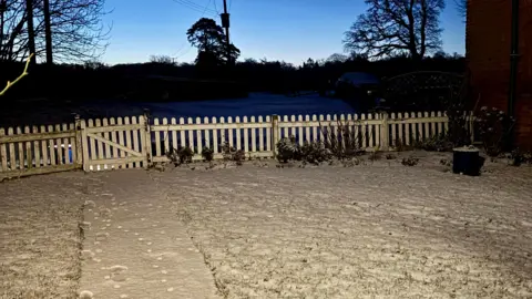 Pauline's view/WEATHERWATCHERS In semi-darkness, a low white fence with a gate runs across the middle of the picture. In front of it, there is a garden covered in snow. Beyond the fence, in the dark, there are the silhouettes of shrubs and trees.