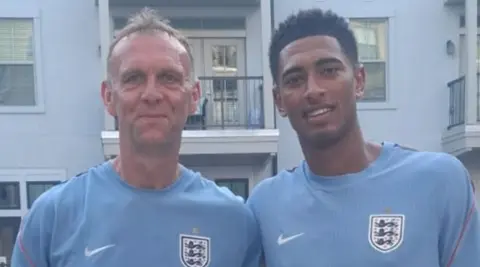 Walton is on the left, next to Bellingham on the right. They are both wearing light blue tops including the coat of arms for the England national team, which features three blue lions. A building is behind the men.