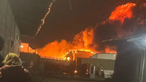 Plumes of smoke, lit orange, rise high in the sky over an industrial site, with lorries visible on the site. The site is fenced off and the back of woman is visible in the bottom-left corner of the image.