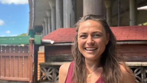 BBC A woman with long brown hair smiling wearing a red vest top standing in front of a wall with a motorway flyover in the background