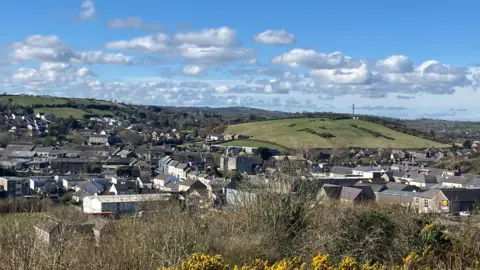 Photo of Ballynahinch taken from a hill. There is gorse in the foreground, then the buildings of the town, and hills in the background. The sky is blue with white clouds.