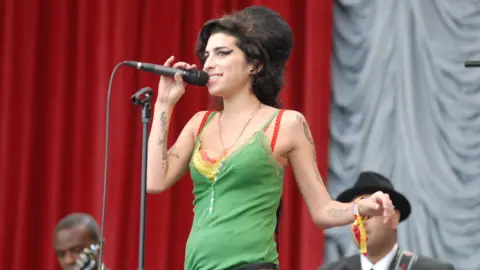 Woman with black hair in green top holds a black microphone while on stage with two musicians behind her