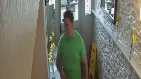 Avon and Somerset Police A CCTV image of a man wearing a bright green polo top walking in a corridor which has white brick walls on either side