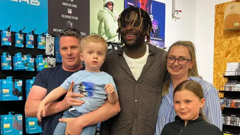 Emma Baugh/BBC Myles Smith poses with the Stockdale family, who consist of a mum, dad, son and daughter. They are inside a HMV shop.
