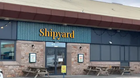 Kate Jennings/BBC The outside of the Shipyard cafe has picnic style tables and chairs with the venue's name in yellow letters against a blue background on the wall.