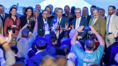 A large crowd with many attendees holding up phones and cameras to photograph a group of people standing on a stage beneath bright lighting and a large screen backdrop
