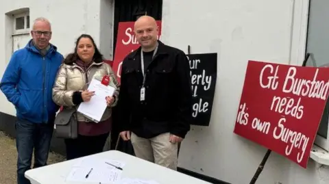 Phil Turner, Justine Norris and Andy Barnes standing outside a white building. They are behind a white table with pens and paper on it. There are black and red placards leaning against the building's external wall with the words: "Gt Burstead needs its own surgery."