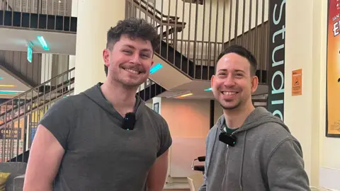 Isabella Verona/BBC Two men standing side-by-side in a bright indoor foyer, with staircases and railings in the background. Both are wearing casual tops and clip-on microphones. A poster is visible on the wall behind them, along with chairs and tables.
