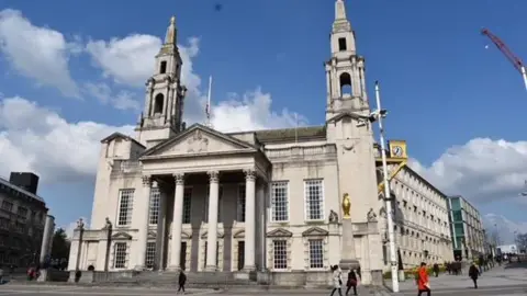 A large, majestic style building supported by three tall columns. On the front of the building are several large windows. 