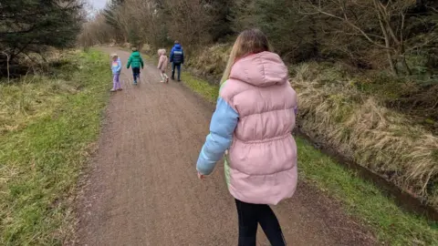 Supplied Laura Earl is walking on a forest pathway with her four children. Everyone is wearing puffy coats and a have their back to the camera. Earl, in the foreground, has long blonde hair and is wearing a pink and blue coat. The children are walking ahead on the pathway that is bordered by trees.