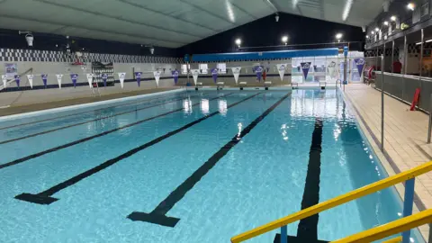 John Devine/BBC six-lane, 25m (82ft) long indoor swimming pool, it has blue and white bunting strung up across the lanes at the deep and shallow ends.
It has new bright white lights fitted in the walls and blue and white ceramic tiles also on the walls. The walkway tiles are a terracotta colour. 