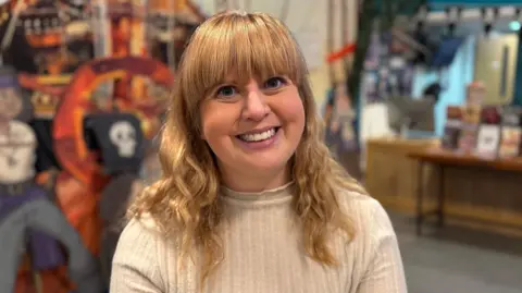 A woman with strawberry blonde wavy hair and a full fringe, smiling and wearing a cream jumper