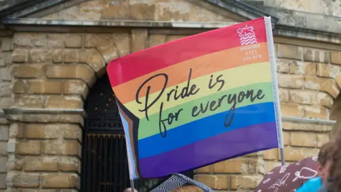 A rainbow flag is waved in front of a historical building in Oxford on 7.6.25. It has the words "Pride is for everyone" and the logo of Oxford City Council, which supported the event.