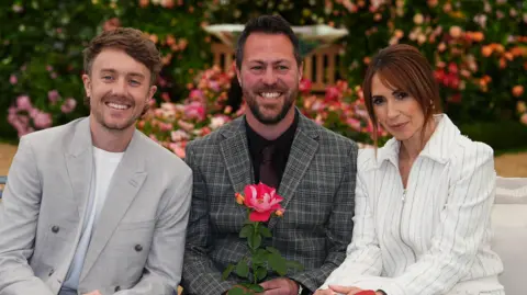 PA Media Roman Kemp, Mat Nicolas and Alex Jones sitting in a row at the Peter Beales RHS Chelsea Flower Shower. On the left is Kemp who is looking directly at the camera and smiling. He is wearing a white T-shirt with a grey blazer over the top. Nicholas is sitting in the middle and is looking directly at the camera and smiling while also holding The One Show rose. He is wearing a black shirt, with a tie and grey blazer over the top. On the right is Jones who is looking at the camera and smiling. She is wearing a white striped co-ord.