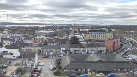 The rooftops of terraced houses and offices spread out near Grays town centre, with the River Thames and pylons visible in the background, set against a cloudy sky.