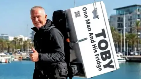 Thomas Judge, a man in his 60s with grey hair wearing a black jacket. He's carrying a fridge on his back. He's clean shaven and is smiling and holding a thumbs up.
