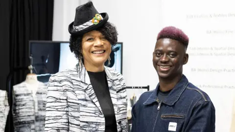 Chi Onwurah wearing a black and white, almost tiger-print suit jacket over a black top. She is wearing a small black hat with a strip of fabric and two orange and green circles. She is smiling into the camera and standing to the right of Lamin Gibba who has pink hair and is wearing a denim jacket. He is also smiling into the camera