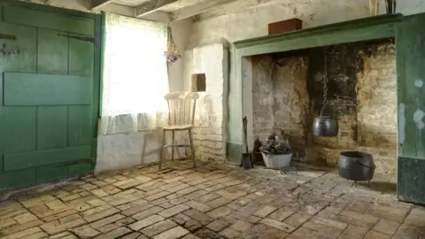 National Trust Images/John Miller A small room has a large fireplace on the right side which has a black cauldron hanging from the ceiling and another one on the floor. There is a wooden chair in the corner of the room which is tiled.