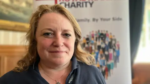 Alice Farrow is blonde and wearing a blue Royal Navy and Royal Marines Charity branded jumper and a silver and black necklace. She is looking at the camera. In the background is a banner displaying the charities branding.