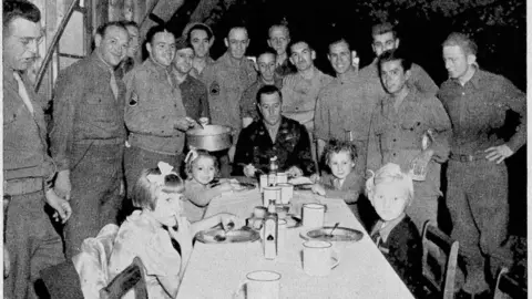 Supplied A black and white photo of children sitting on a long rectangular table. They have food in front of them and look happy. Behind them are US airmen in uniform, smiling at the camera.