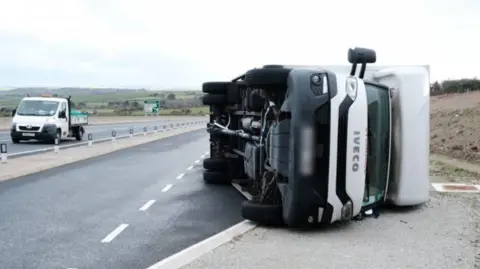 PA Media The picture shows a large white Iveco lorry lying on its side on the edge of a road. The vehicle appears to have overturned, with its wheels facing sideways and its undercarriage fully visible. The cab is intact, but the position suggests a serious incident, likely caused by strong winds or loss of control. The lorry is resting partly on the tarmac and partly on a gravel shoulder