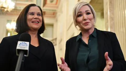 Getty Images Mary Lou McDonald, left, with shoulder length brown hair wearing a black jacket standing in Stormont in front of a microphone.  Michelle O'Neill, with short blonde hair, is standing beside her wearing a dark grey top and black jacket.